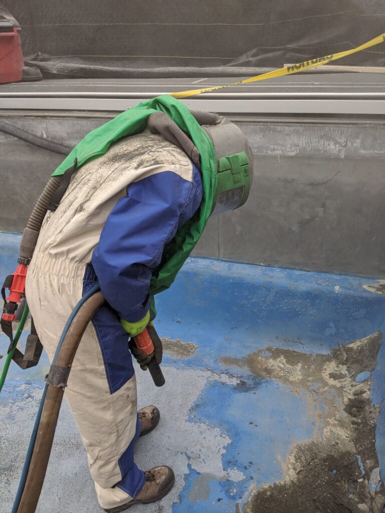 Worker using slurry blasting equipment for surface preparation, wearing protective gear, in an industrial setting, demonstrating dust suppression and cleaning process.