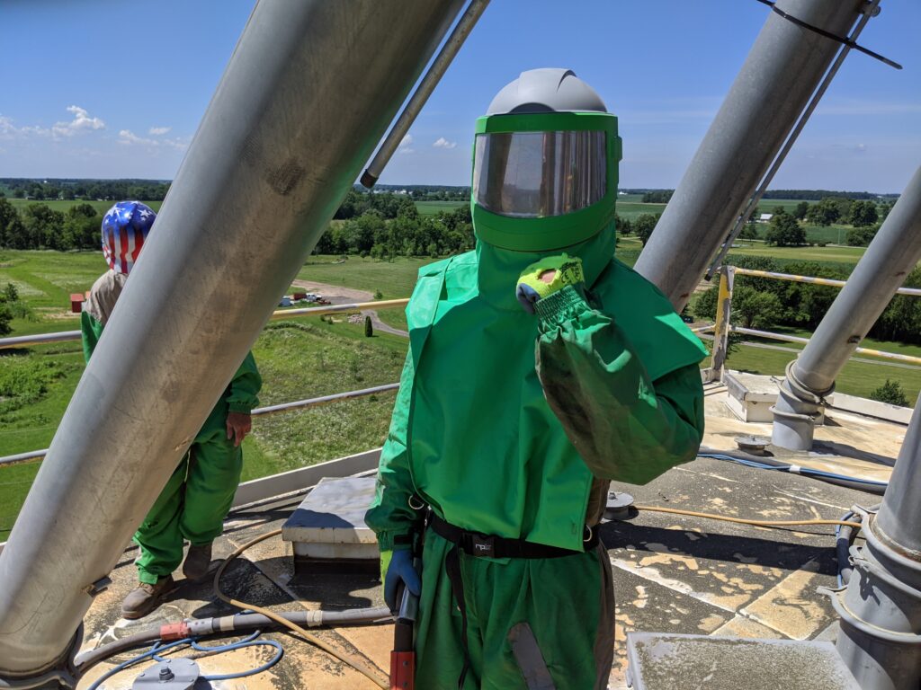 Worker in green protective gear demonstrating dry ice blasting technology on industrial site, with clear skies and green landscape in background.