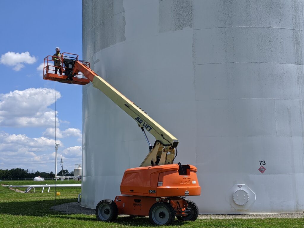 Worker in a lift performing maintenance on a large industrial storage tank, emphasizing tank coatings and linings for corrosion protection.