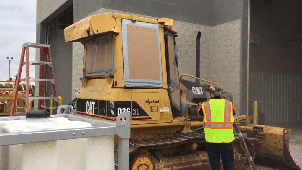 Worker in safety gear cleaning a CAT D3K XL bulldozer with a hose, showcasing maintenance procedures relevant to commercial cleaning assessments.